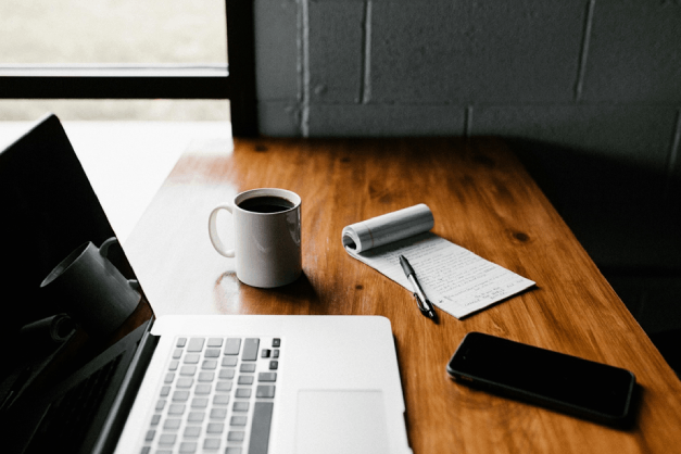 Computador sobre una mesa, con una libreta , una taza y un celular 